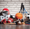 Sports equipment laid out on a floor against a brick wall: helmets, pads, gloves, and balls from multiple sports (football, basketball, soccer).